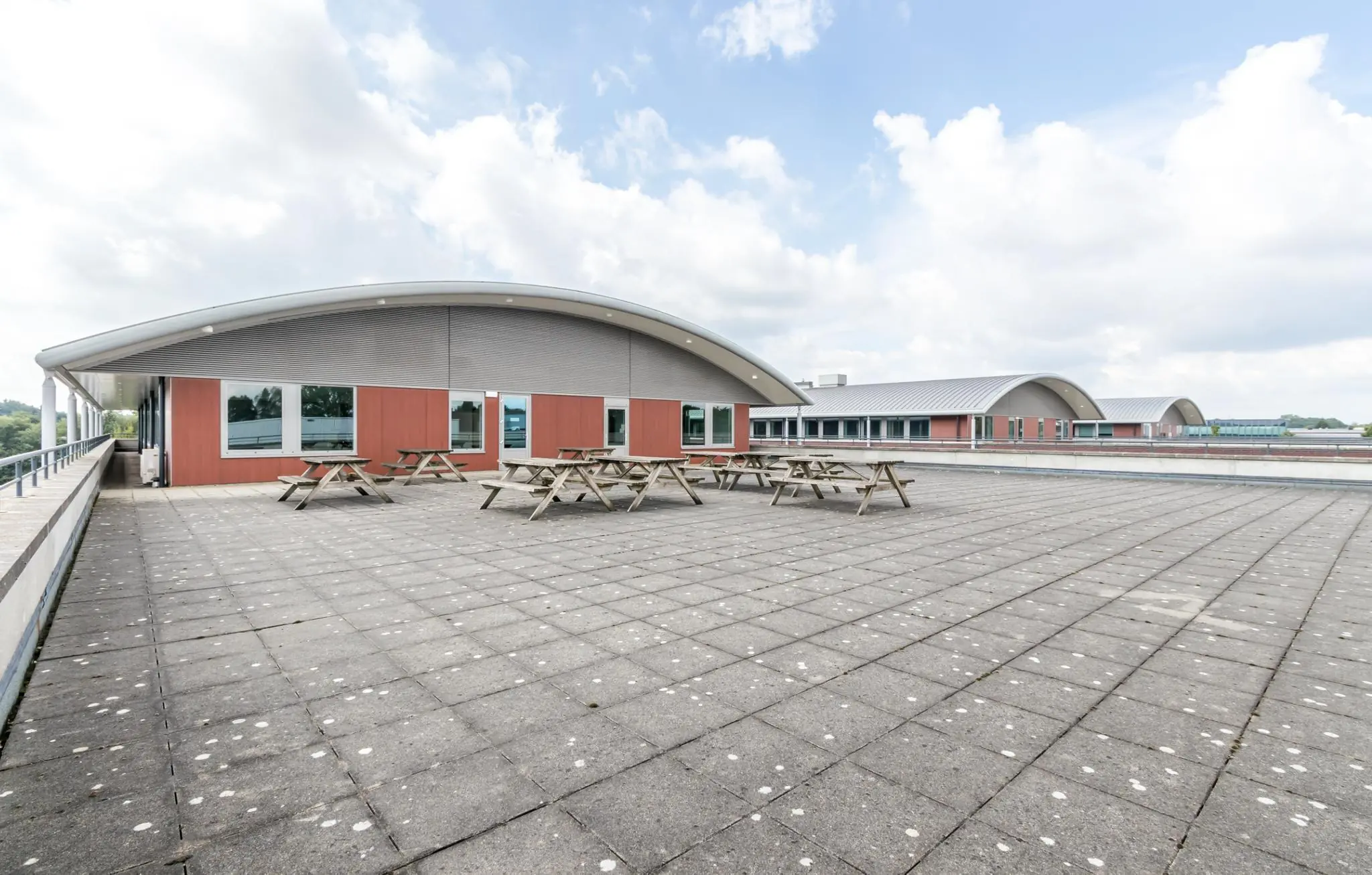Ruim dakterras bij De Wel met meerdere picknicktafels en uitzicht op moderne schoolgebouwen onder een deels bewolkte hemel.